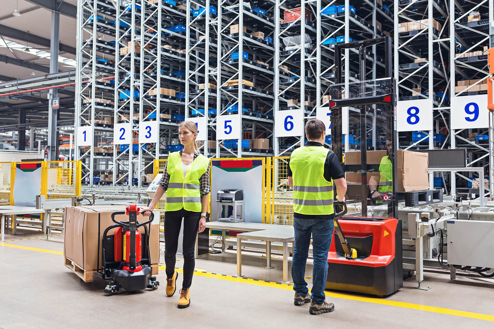 Storehouse employees in uniform standing near pallet truck and forklift in modern automatic warehouse.Boxes are on the shelves of the warehouse. Warehousing, machinery concept. Logistics in stock